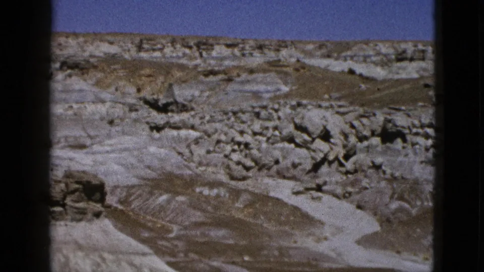 1953 Arizona home-movie frame showing eroded desert terrain, layered rock, sparse scrub, and clear blue sky.