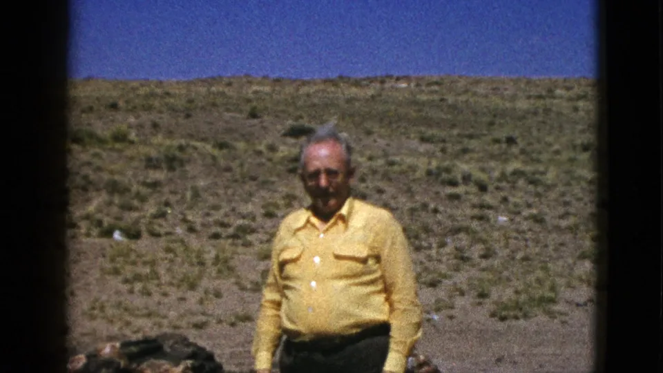 An older man in a yellow button-up shirt faces the camera in a sunny Arizona desert landscape, filmed on 8mm in 1953.
