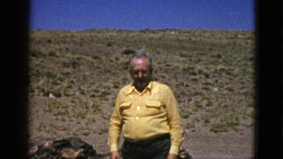 The man remains facing the camera in the Arizona desert, with low scrub, rocks, and a clear blue sky behind him.