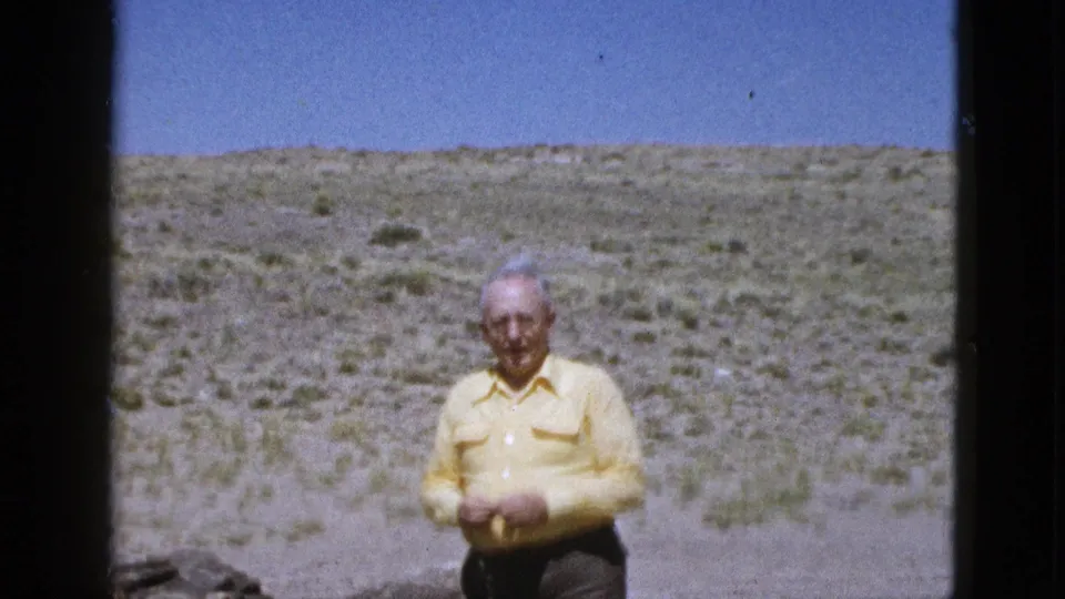 The man is framed slightly smaller in the Arizona desert, standing in bright sun with sparse scrubland stretching behind him.