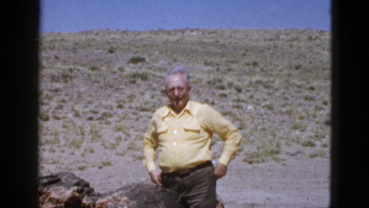 1953 Arizona home-movie frame showing an elderly man in a yellow shirt and dark pants standing before scrubby desert hills.