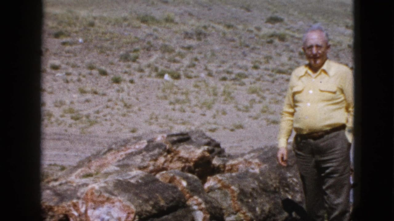 Elderly man standing beside weathered rock in an arid landscape.