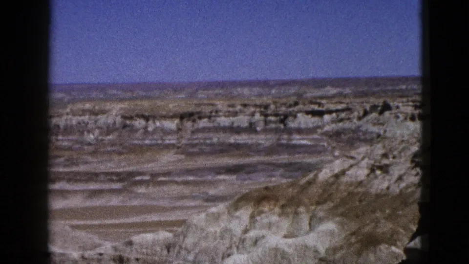 1953 Arizona 8mm frame with pale layered desert ridges, dry foreground slopes, and a clear blue sky.