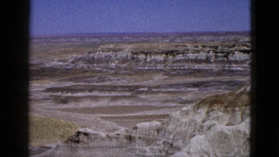 1953 Arizona 8mm frame with a rocky foreground slope, dry basin, distant ridge, and clear blue sky.