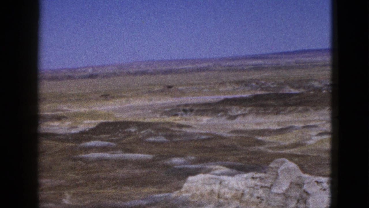 1953 Arizona 8mm frame with rolling arid ground, pale outcrops, and a distant ridge line under clear blue sky.