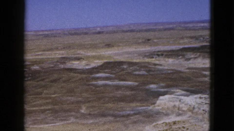 1953 Arizona 8mm frame capturing a wide arid vista, pale lower-right rocks, and layered desert flats under a blue sky.