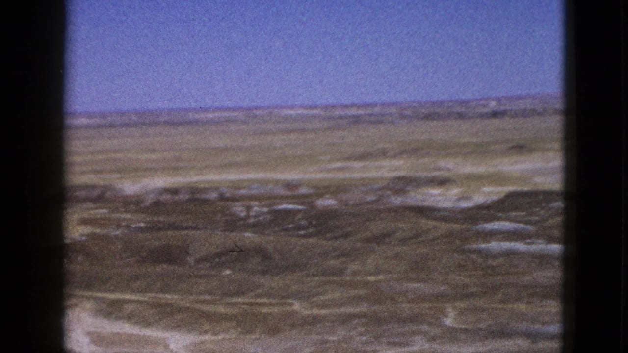1953 Arizona desert terrain with low ridges, pale washes, and blue sky seen through dark 8mm frame borders.
