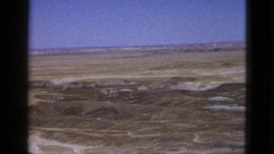 1953 Arizona desert terrain with low ridges, pale washes, and blue sky seen through dark 8mm frame borders.