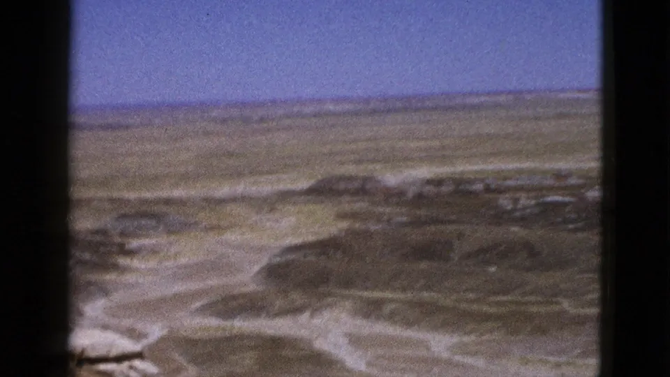 1953 Arizona home-movie frame showing an arid desert panorama with low mesas, pale rock layers, and open blue sky.