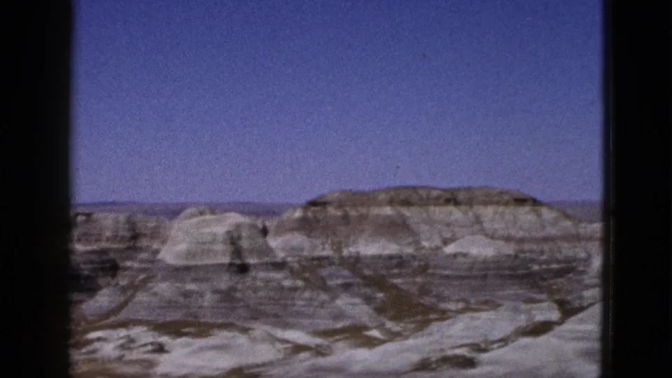 1953 Arizona frame with banded desert hills, pale eroded rock faces, sparse ground cover, and a clear blue sky.