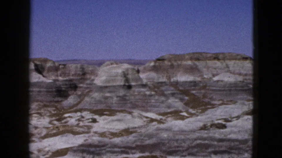 1953 Arizona 8mm view of arid terrain with layered rock bands, rounded pale slopes, and sparse desert surface.