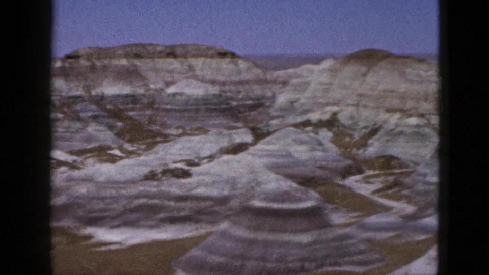 Arizona desert badlands appear in a mid-century 8mm frame, showing stratified grey and tan hills with dry drainage lines.