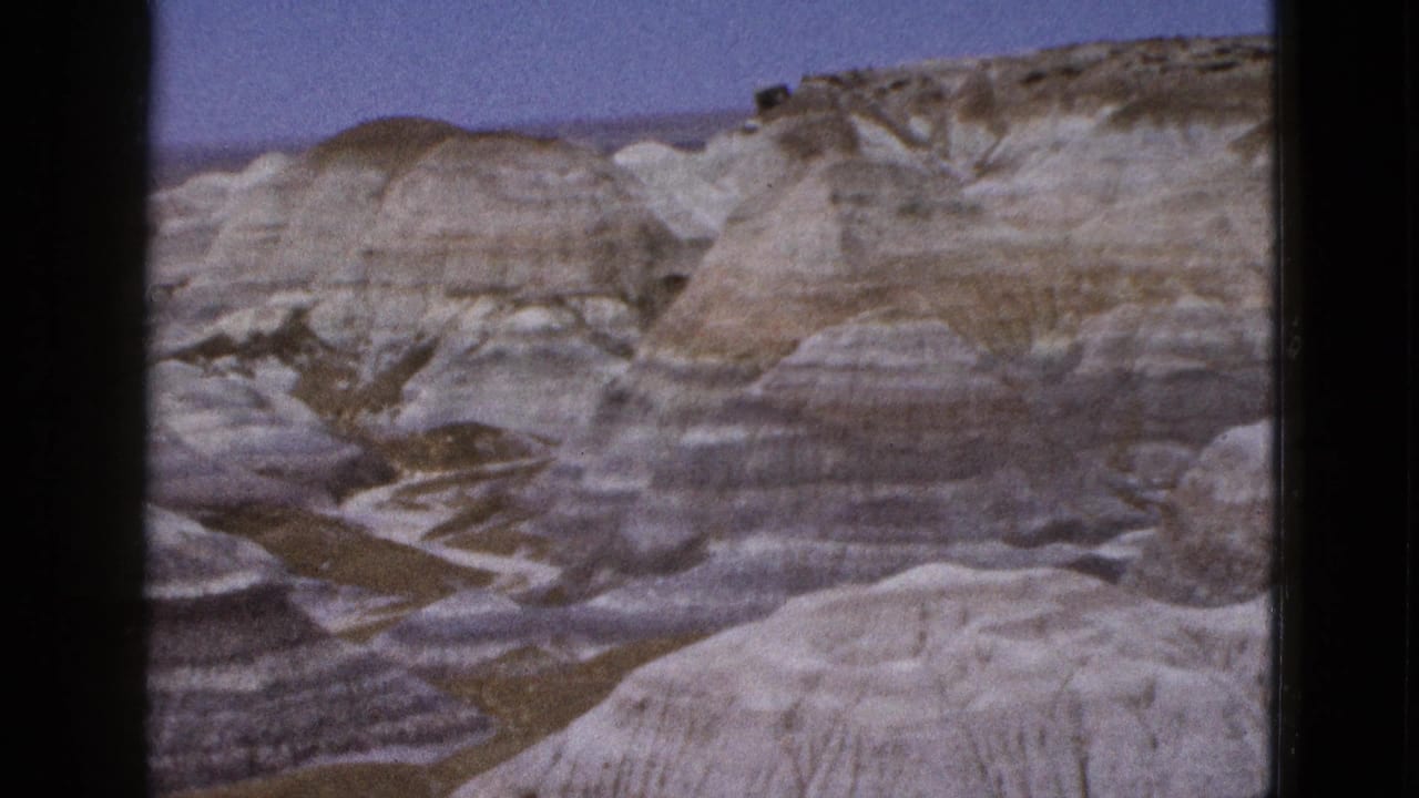 Banded desert rock formations below a pale blue sky with dark 8mm frame edges.