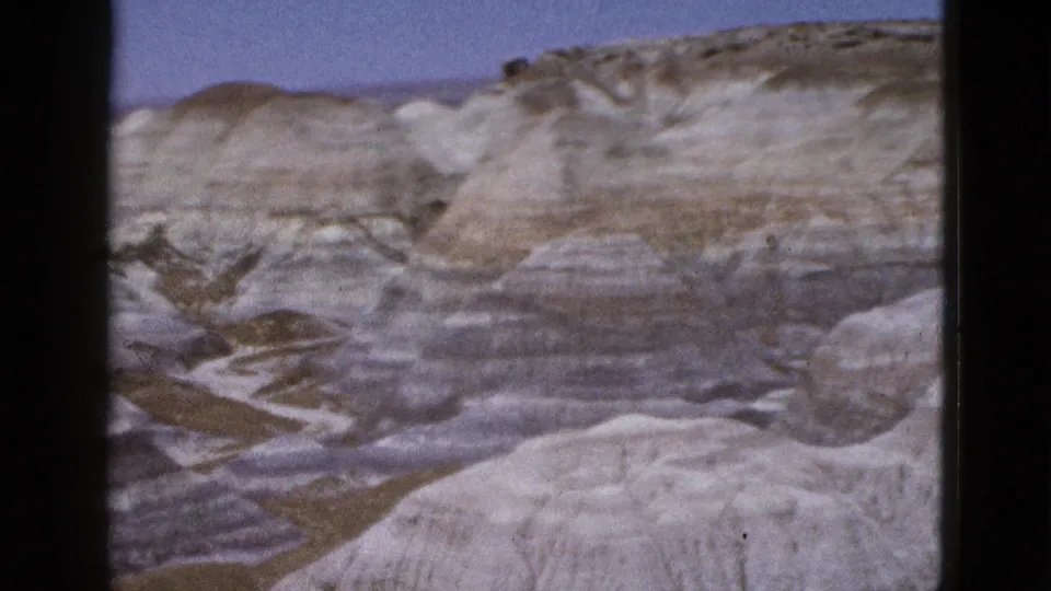 1953 Arizona 8mm frame showing stratified desert hills, a foreground pale slope, gullies, and sky.