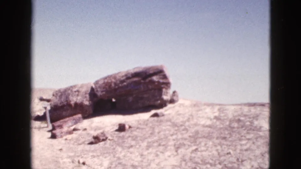 1953 Arizona 8mm frame showing a large weathered boulder resting on barren pale ground under a clear blue sky.