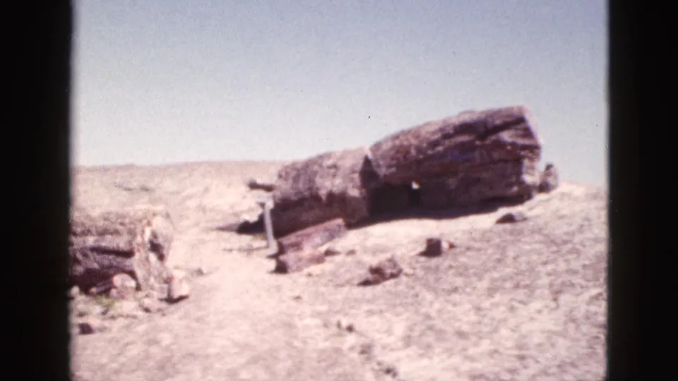 1953 Arizona 8mm frame with a prominent log-like rock formation crossing a dry, lightly scrubbed desert slope under open sky.
