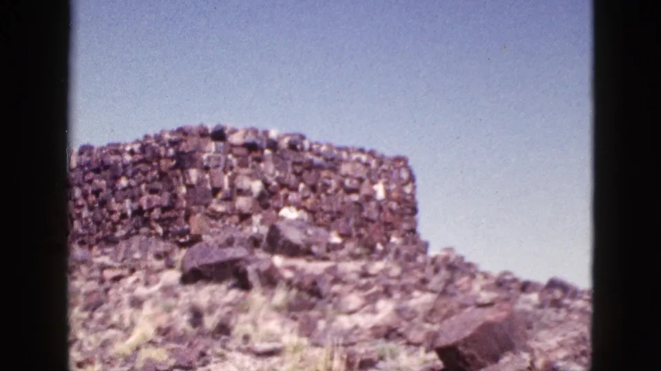 1953 Arizona 8mm frame showing a rough stone ruin wall on rocky desert ground under a clear blue sky.