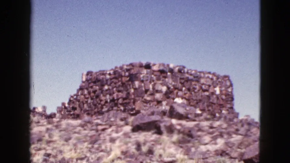 1953 Arizona home-movie frame of a rough stone ruin wall viewed from below on a rocky desert rise.