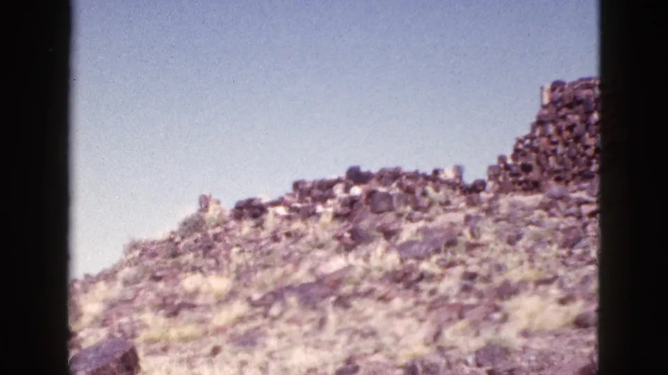1953 Arizona 8mm frame showing a rocky desert slope with sparse grass and the edge of a stone ruin near the ridge.