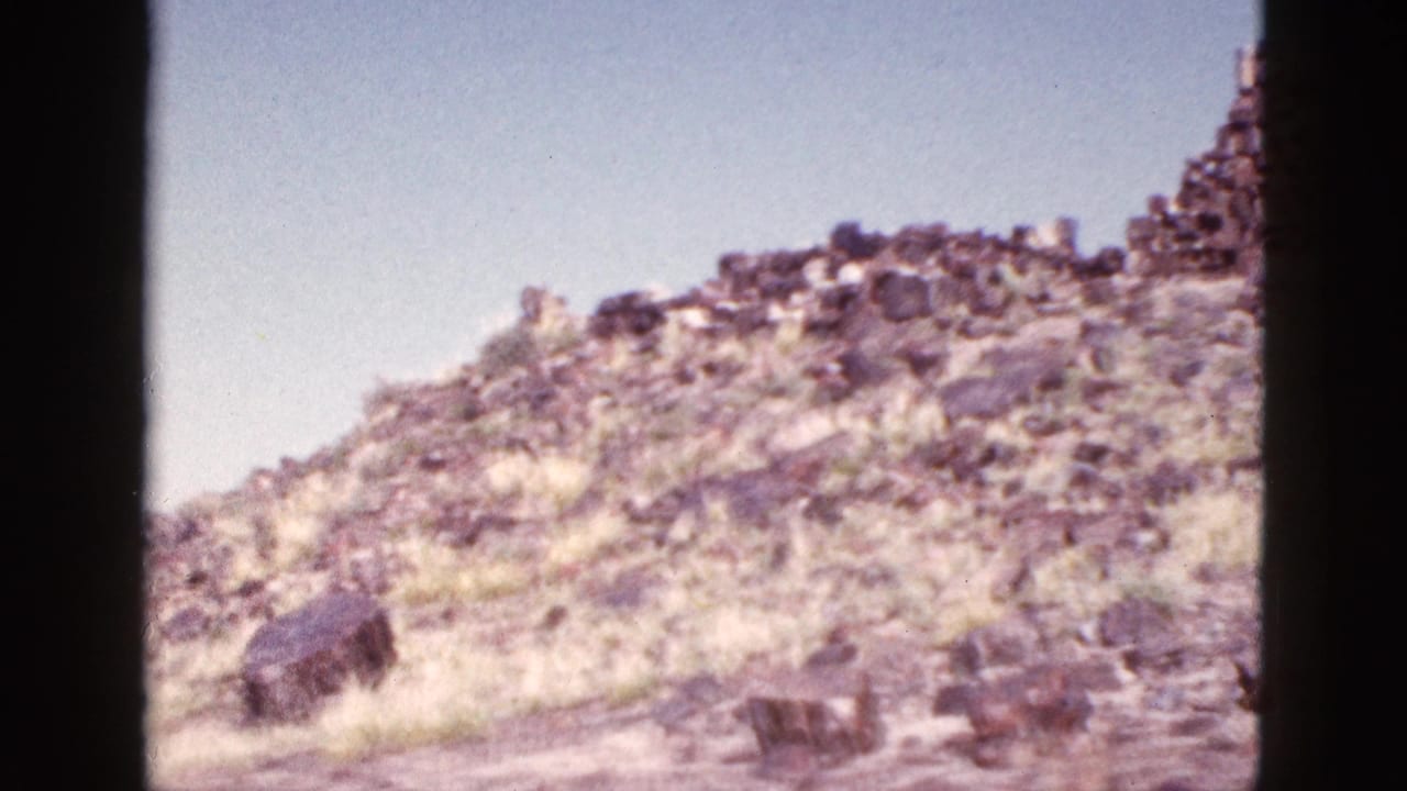 1953 Arizona 8mm frame showing a sunlit rocky slope, sparse dry grass, and a low stone structure beneath blue sky.