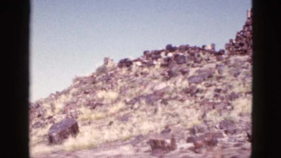 1953 Arizona 8mm frame showing a sunlit rocky slope, sparse dry grass, and a low stone structure beneath blue sky.