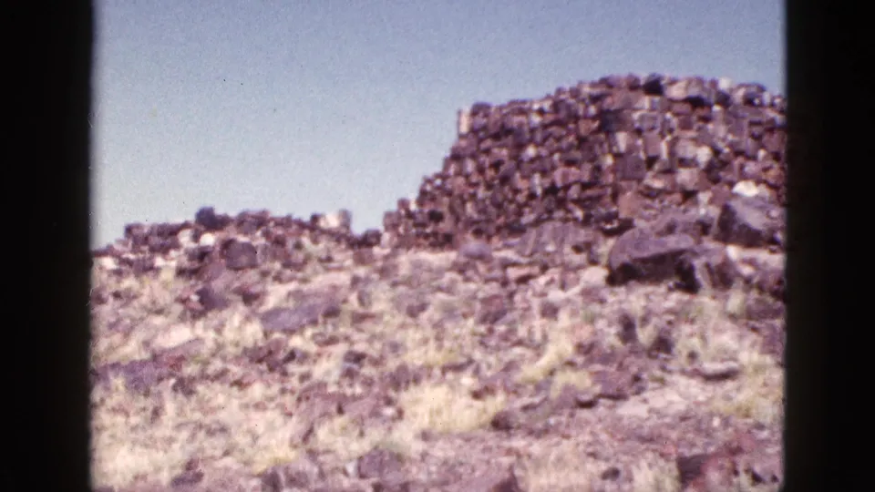 1953 Arizona 8mm frame showing sunlit desert rocks, pale grass, and a rounded section of stacked stonework.