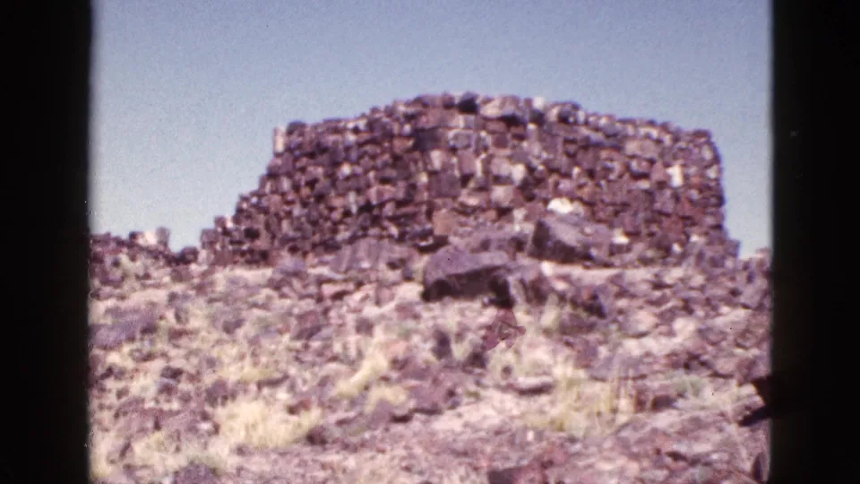 1953 Arizona home-movie frame showing a rough stacked stone structure, boulders, dry grass, and bright blue sky.