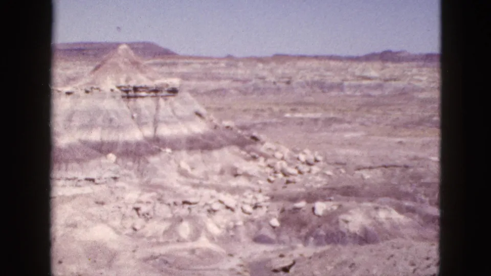 1953 Arizona home-movie frame with pale eroded rock mounds, stony slopes, distant ridges, and open blue sky.