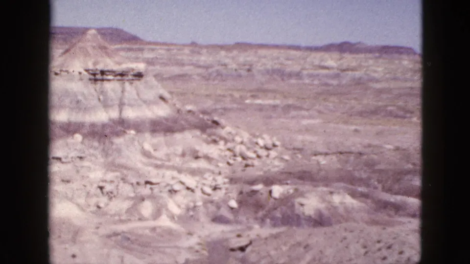 1953 Arizona 8mm frame showing an eroded canyon mound, scattered rocks, open desert floor, and low mesas in the distance.