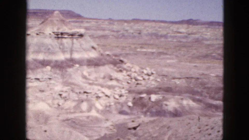 1953 Arizona home-movie frame of pale badlands, eroded mounds, loose stones, distant mesas, and bright blue sky.