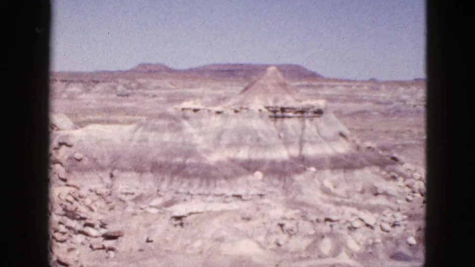 1953 Arizona 8mm frame of eroded desert rock, scattered stones, and a small conical butte in the distance.