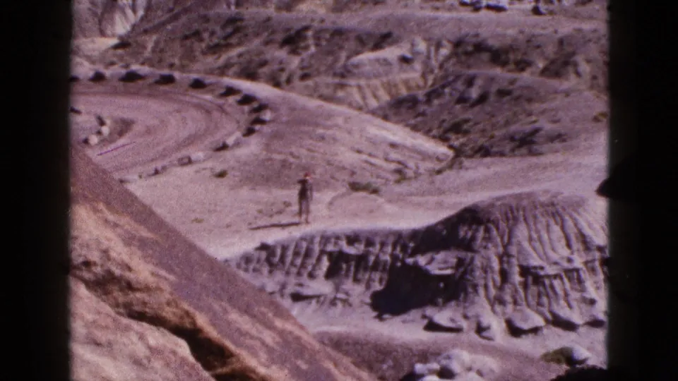 1953 Arizona 8mm frame looking across eroded desert hills, with a distant man standing near the road cut.