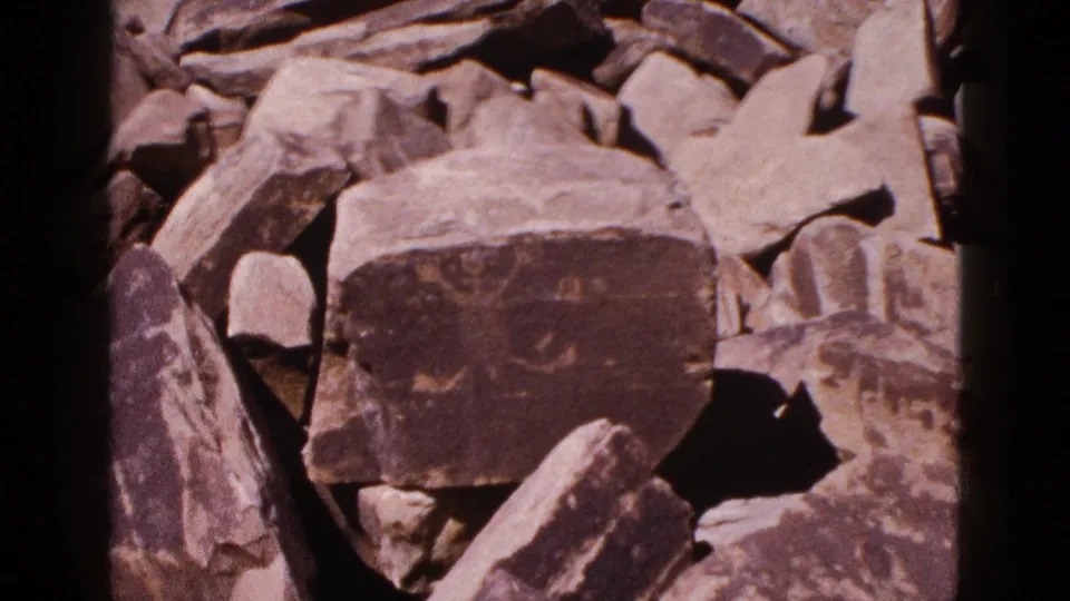 1953 Arizona 8mm frame showing faint petroglyph markings on a dark boulder surrounded by broken stone slabs.