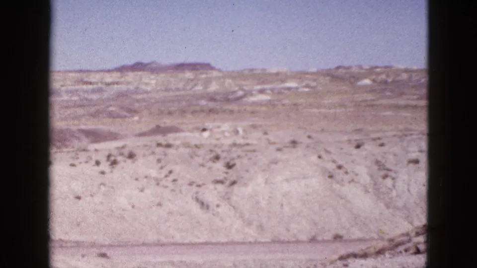 1953 Arizona desert panorama with rolling pale ground, scattered shrubs, low rocky hills, and a clear blue sky.