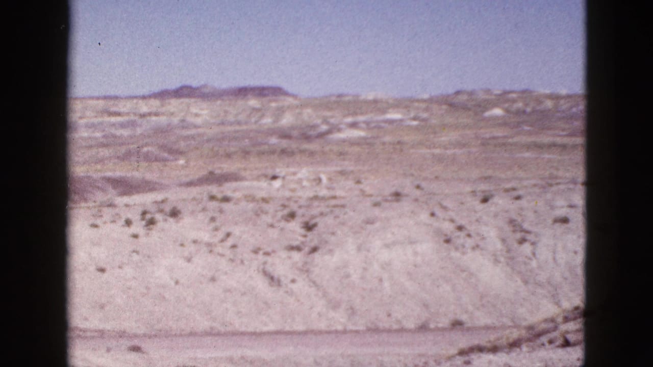 1953 8mm Arizona home-movie frame showing open desert terrain, low vegetation, distant mesas, and a clear blue sky.