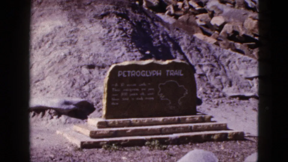 1953 Arizona 8mm frame showing a Petroglyph Trail stone marker on a stepped base with rocks and desert ground behind it.