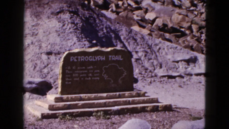 1953 Arizona home-movie frame of the Petroglyph Trail marker, showing engraved lettering, a map outline, steps, and surrounding rocks.