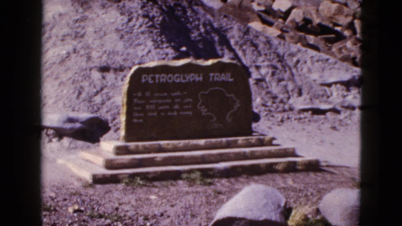 Dark Petroglyph Trail marker with lettering and map outline beside a rocky slope.
