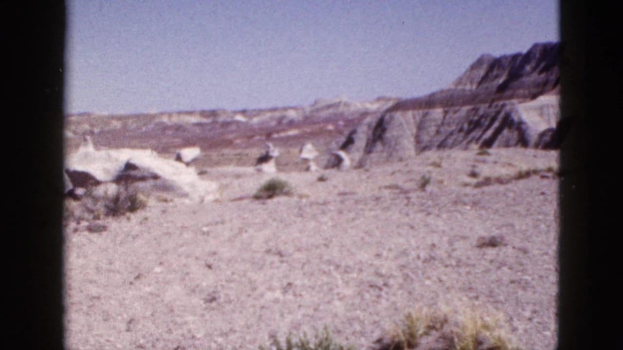 1953 Arizona 8mm frame showing open desert ground, sparse brush, pale eroded mounds, layered cliffs, and blue sky.