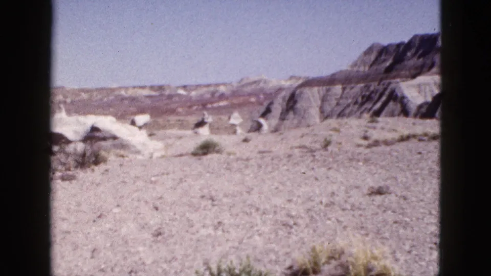 1953 Arizona 8mm frame with gravelly desert foreground, sparse vegetation, pale eroded rocks, and distant mesas.