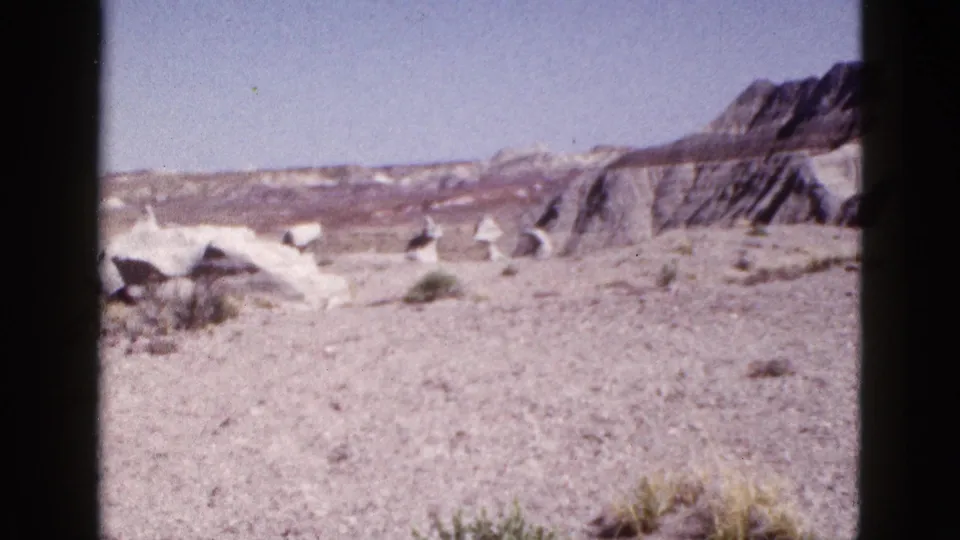 1953 Arizona home-movie still showing a wide arid view with low shrubs, pale rock mounds, and layered cliffs.
