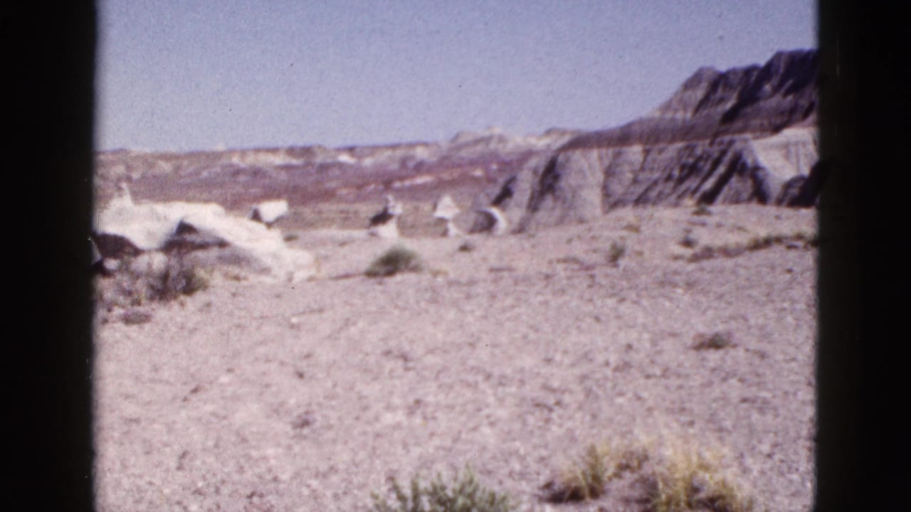 1953 Arizona 8mm frame showing barren desert ground, scattered rocks, scrub vegetation, and distant mesa formations.
