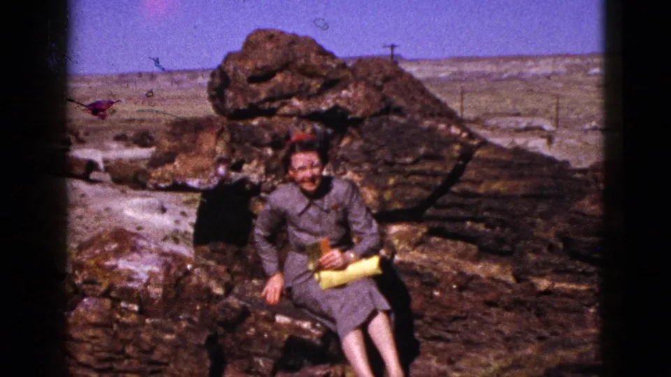 1953 Arizona 8mm frame of an older woman posing on a large dark boulder, wearing a gray dress and red head covering while holding a yellow purse.