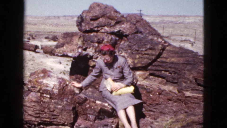1953 Arizona home-movie frame showing a woman seated on a weathered boulder, holding a yellow purse against a rocky desert backdrop.