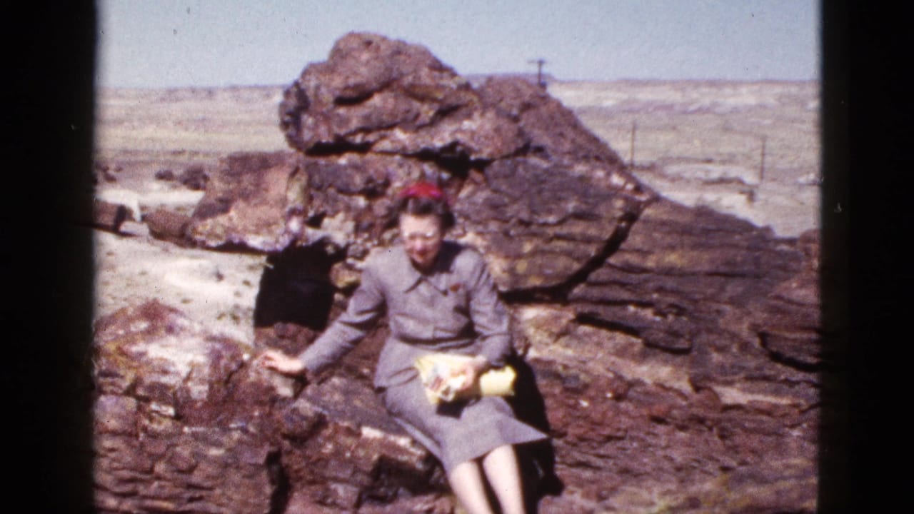 Woman in a gray dress and red hat sits on desert rocks holding yellow papers.