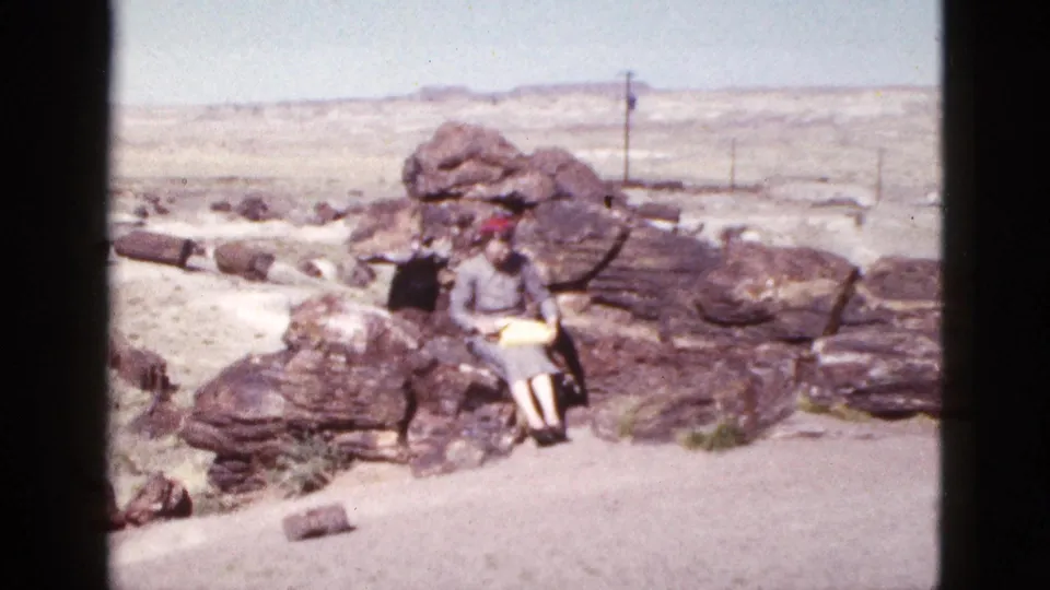 A wider 1953 Arizona view shows the woman sitting among large rocks with desert terrain and utility poles behind her.