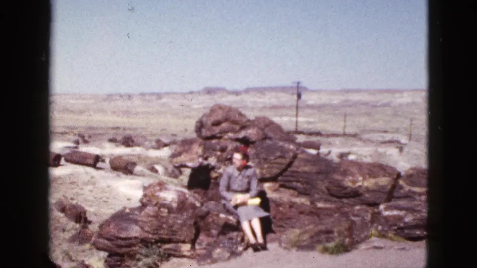 1953 Arizona home-movie still of a woman in a hat seated on a rock formation in a windy-looking desert setting.