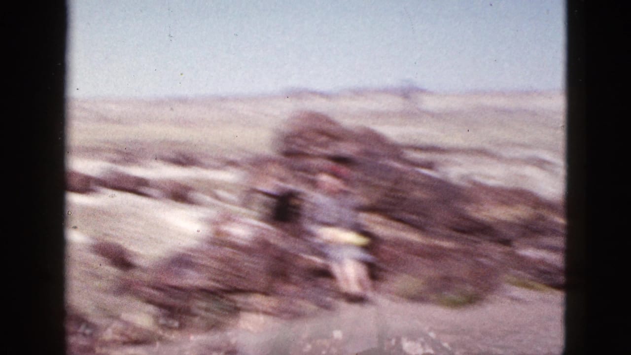 Soft, motion-blurred 1953 Arizona desert home-movie frame with rocks, open terrain, and a distant person in yellow.