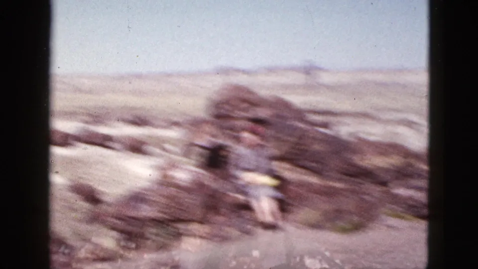Soft, motion-blurred 1953 Arizona desert home-movie frame with rocks, open terrain, and a distant person in yellow.