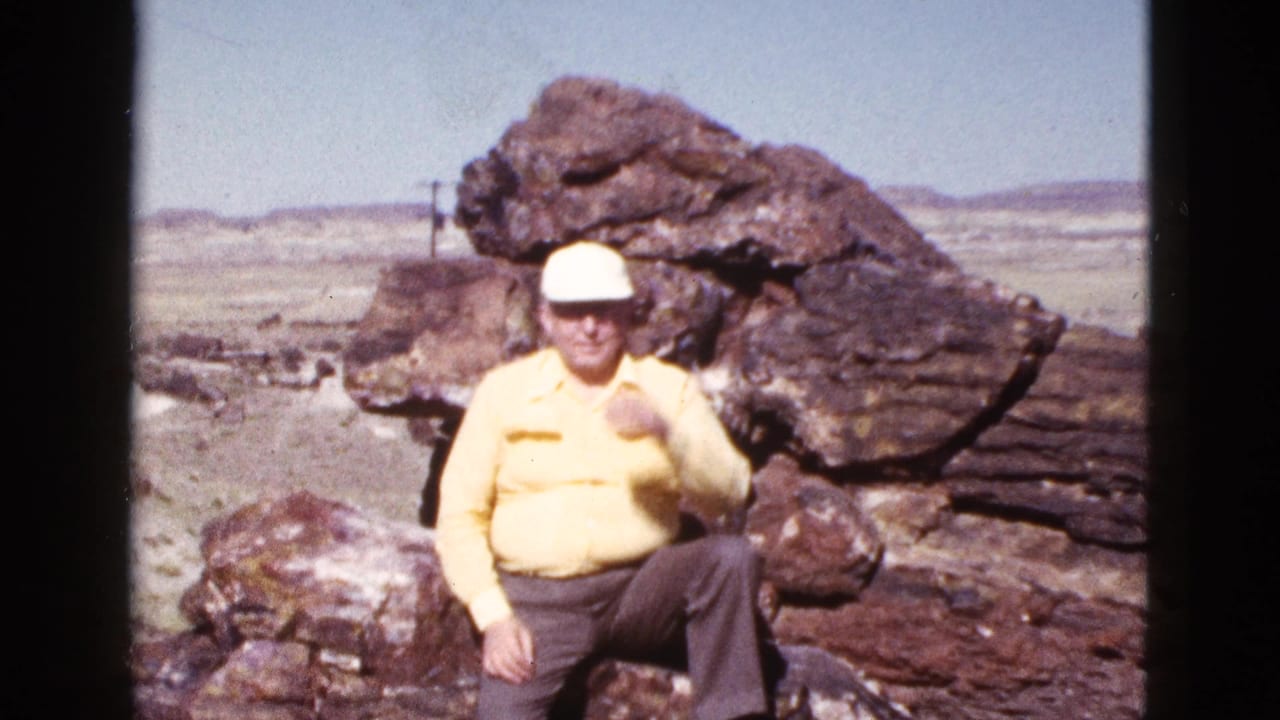 Older man in a yellow shirt and white cap sits against large rocks in the Arizona desert, facing the camera.
